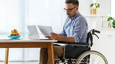 Applied Cloud A man in a wheelchair, sitting at a desk in front of his laptop while holding some papers