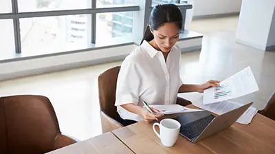 Download Services  Woman dressed in business casual attire sitting at a table with a coffee mug and laptop while looking over papers.