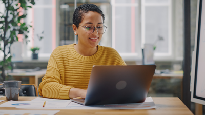 Looking Back, Looking Ahead: Accomplishments, Reflections & Gratitude A woman in an orange sweater sitting at her desk and smiling while typing on her laptop.
