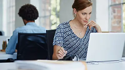 Applied Policy Works  A woman sitting at a desk and using a laptop while taking notes. A man is sitting at a desk in the background  while working.