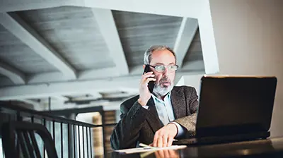 Church West Customer Success Man dressed in business attire sitting at table and holding phone to ear while looking at laptop.