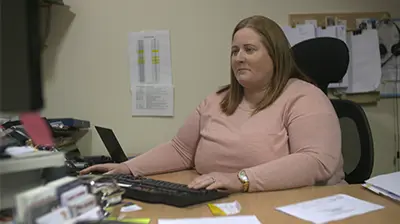 Gollogly Insurances Ltd enables mobility and scalability with the cloud Woman in casual attire sitting behind a desk in an office, she is looking at a computer screen in front of her with her hand on a keyboard.