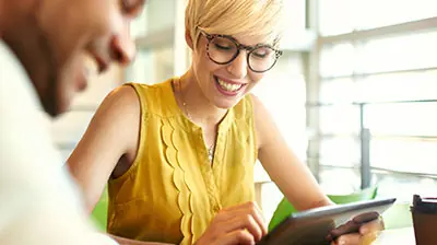 The Digital Age of Insurance Smiling woman in casual attire with glasses looking down at a tablet in her hands, there is a man sitting in front of her to the left of the image, also smiling.