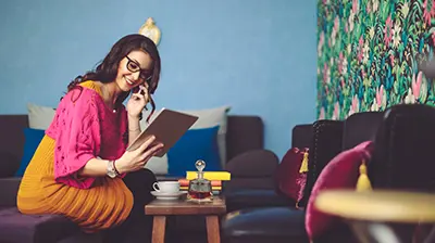 7 Fast Facts for Insuring the Digital Generation Smiling woman in brightly colored clothing, holding a cell phone to her ear while looking down at a tablet device in her left hand. She’s sitting on a couch with a coffee table in front of her.
