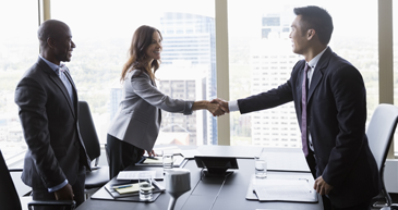 Industry Partners A group of people in business attire standing at a table in an office, a man and woman are shaking hands across the table.