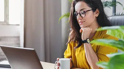 Taking Applied Epic to the Browser A woman sitting at her desk while using her laptop. She is holding a mug in one hand and resting her other hand underneath her chin.