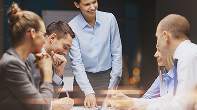 Applied Partnership Overview A businesswoman standing at the head of the table speaking while four colleagues are sitting around the table while listening