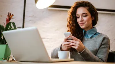 Applied Digital Agency Annual Report A woman in business attire sitting at a desk in front of an open laptop, while looking at a smartphone that she is holding with both of her hands.